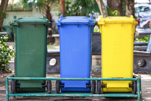The image displays three large, wheeled rubbish bins positioned side by side outdoors on a paved area, likely on a residential street or communal parking space. The bin on the left is green with a textured, matte finish, and is slightly taller than the others; it features a closed lid and a durable plastic body. The middle bin is blue, with a similarly textured surface and a closed lid, appearing to be used for general waste or recycling. The rightmost bin is yellow, also with a closed lid and a smooth, slightly glossy surface, which could be designated for recyclable materials. All three bins are positioned on a metal trolley with black rubber wheels, which supports easy transportation and waste collection. In the background, there are several trees with brown trunks and green foliage, indicating a park or garden environment, and some parked vehicles are visible beyond the bins. The lighting suggests daytime with natural sunlight illuminating the scene. The overall setting reflects a typical outdoor waste collection arrangement, suitable for commercial or community waste removal services by companies like Commercial Waste Forest Hill, serving local areas in Forest Hill, London, with postcode areas inferred from the context.