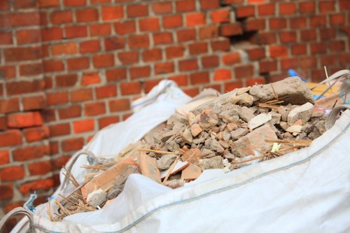 A large white woven polypropylene sack filled with a mixture of red and beige broken bricks, concrete debris, and small pieces of tile, situated outdoors on a paved surface in front of a red brick wall. The debris appears to be construction or renovation waste, with some of the bricks and concrete fragments showing rough texture and weathered surfaces. The sack is partially open at the top, revealing the contents inside, and is positioned on the ground near a building, possibly in preparation for rubbish removal service. The environment suggests a residential or commercial property in Forest Hill, with the presence of construction waste awaiting collection by a company like Commercial Waste Forest Hill, which specializes in rubbish removal services.