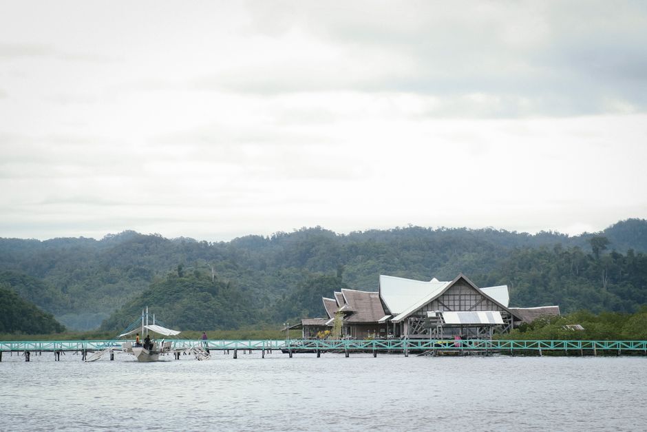 The image depicts a waterfront scene featuring a wooden structure with traditional-style architectural elements, including a steeply pitched roof and timber framing, situated on stilts above calm wate