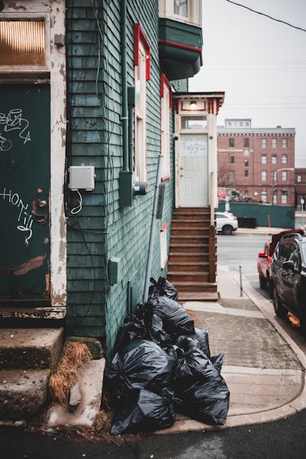 A collection of several black garbage bags piled against the green wooden exterior wall of a building in an urban setting. The bags appear to be full and are placed on a small concrete platform beside